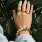 A hand wearing three jade rings and a Gradient Green Nephrite Jade Bracelet—natural celadon-to-olive green stone beads for fashion & energy—is held before green leafy plants. The person wears a light blue sweater.