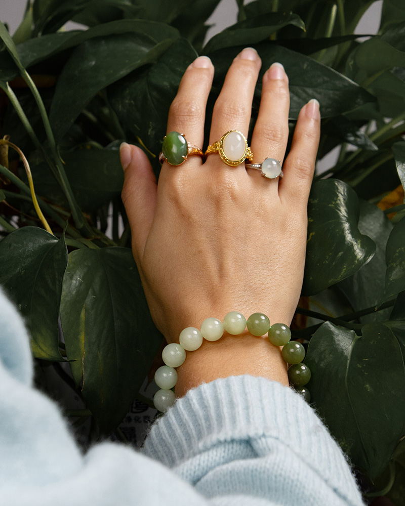 A hand wearing three jade rings and a Gradient Green Nephrite Jade Bracelet—natural celadon-to-olive green stone beads for fashion & energy—is held before green leafy plants. The person wears a light blue sweater.