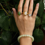 A hand adorned with the Handmade Jade Bracelet – Natural Nephrite Stone Jewelry rests over green leafy plants, complemented by three gemstone rings in green, white, and light gray.