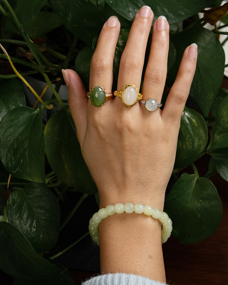 LYU_4218 A hand adorned with the Handmade Jade Bracelet – Natural Nephrite Stone Jewelry rests over green leafy plants, complemented by three gemstone rings in green, white, and light gray.