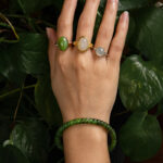 A hand wearing the Natural Green Nephrite Jade Bracelet with three gold gemstone rings—featuring oval green, white, and pale gray stones—rests on leafy plants. The person is dressed in a light blue sweater.