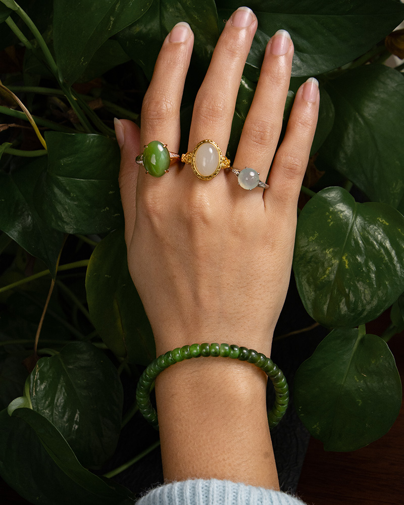 LYU_4219 A hand wearing the Natural Green Nephrite Jade Bracelet with three gold gemstone rings—featuring oval green, white, and pale gray stones—rests on leafy plants. The person is dressed in a light blue sweater.