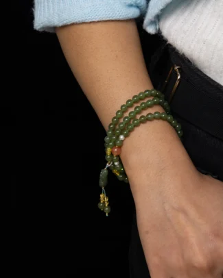 A person wearing the Grayish Green Nephrite Jade Bracelet – Spherical Beads Multi-Wrap with Carved Focal, wrapped around their wrist, paired with a white sweater and dark pants, standing against a black background.