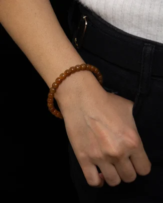 A person wearing the Cognac Brown Nephrite Jade Bracelet – Drum Beads, Gloss-Polished Strand on their wrist, dressed in a white top and black pants with a belt, stands against a black background.