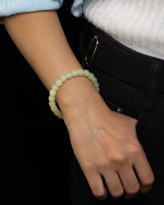 A person wears the Pale Cream Nephrite Jade Bracelet – Drum Beads, Soft-Gloss Finish on their wrist, paired with a white sweater and dark pants, their hand relaxed by their side against a black background.