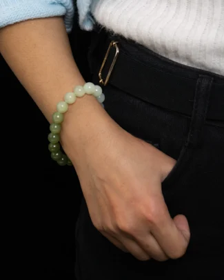 A person wearing the White-to-Olive Nephrite Jade Bracelet – Spherical Beads, Gradient Strand has their hand in the pocket of black pants, with a white ribbed top and light blue sweater visible.