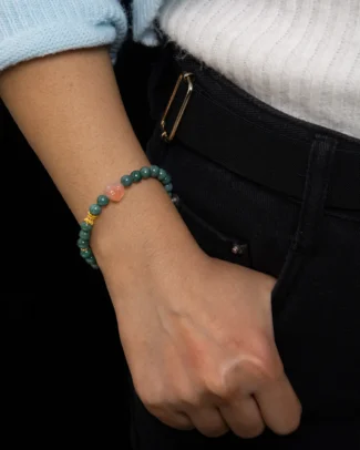 A person wearing the Jadeite Bead Bracelet — Deep Green Single-Wrap with Quartzite Carved Centerpiece has their left hand tucked into the pocket of black jeans. They are dressed in a white ribbed sweater with light blue sleeves.