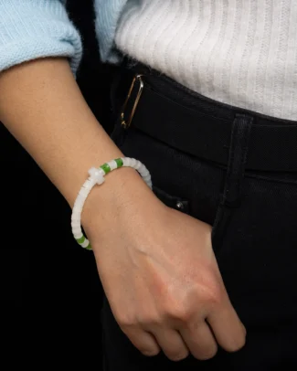 A person wears black pants, a white ribbed shirt, and a pale blue sweater. Their hand rests in their pocket, revealing the Mist-White Nephrite Jade Bracelet – featuring carved disc beads with vivid green accents and a cross charm.