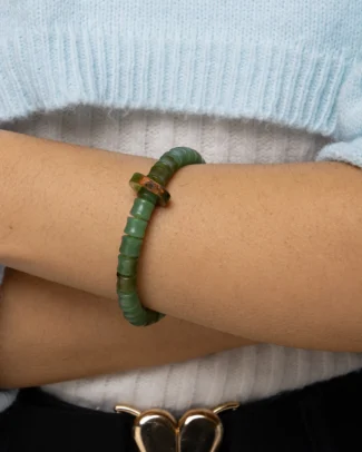 A person wearing a light blue sweater and a Green Nephrite Jade Bracelet featuring carved ring beads with a brownish-red accent, arms crossed over their torso. The outfit includes a belt with a gold heart-shaped buckle.