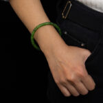 A person wears a Natural Green Nephrite Jade Bracelet—handmade with polished round beads—for men and women, with their hand in the pocket of black pants, dressed in a white sweater and light blue sleeve against a black background.