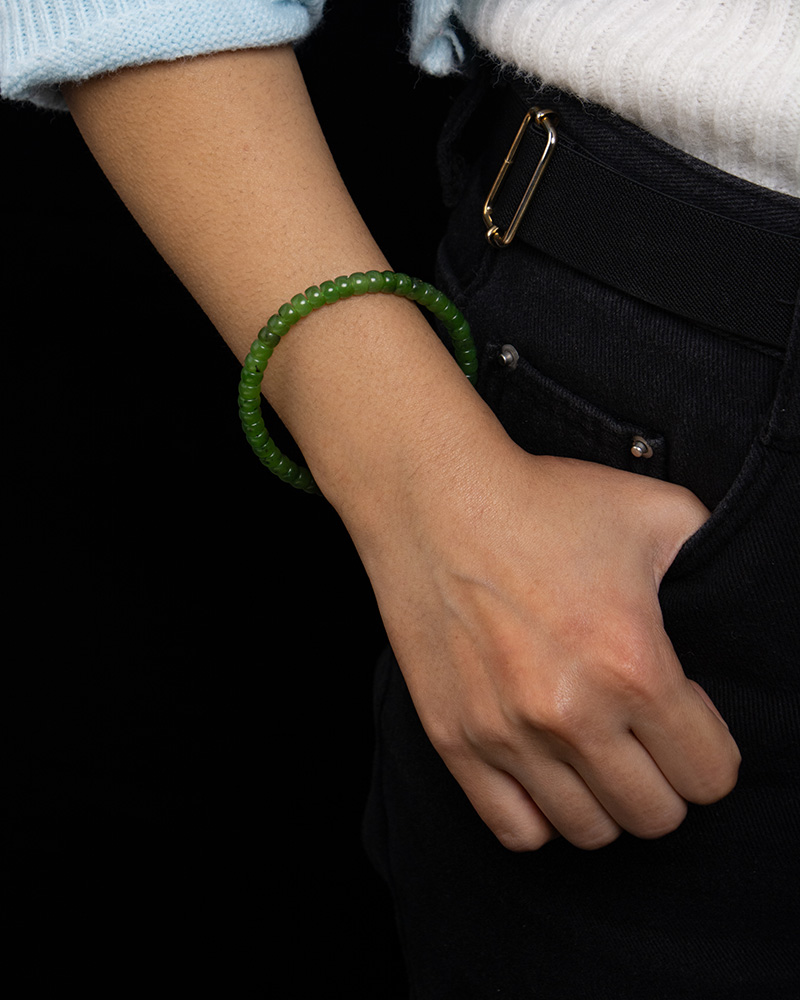 LYU_4248 A person wears a Natural Green Nephrite Jade Bracelet—handmade with polished round beads—for men and women, with their hand in the pocket of black pants, dressed in a white sweater and light blue sleeve against a black background.