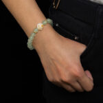 A person in dark pants and a white top has a hand in their pocket, displaying the Mint Green Aventurine Quartz Bracelet with round beads and a cat paw charm.