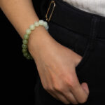 A person wears the Gradient Green Nephrite Jade Bracelet—celadon-to-olive green natural stone beads—resting their hand in a dark jeans pocket, with a white sweater at the wrist.