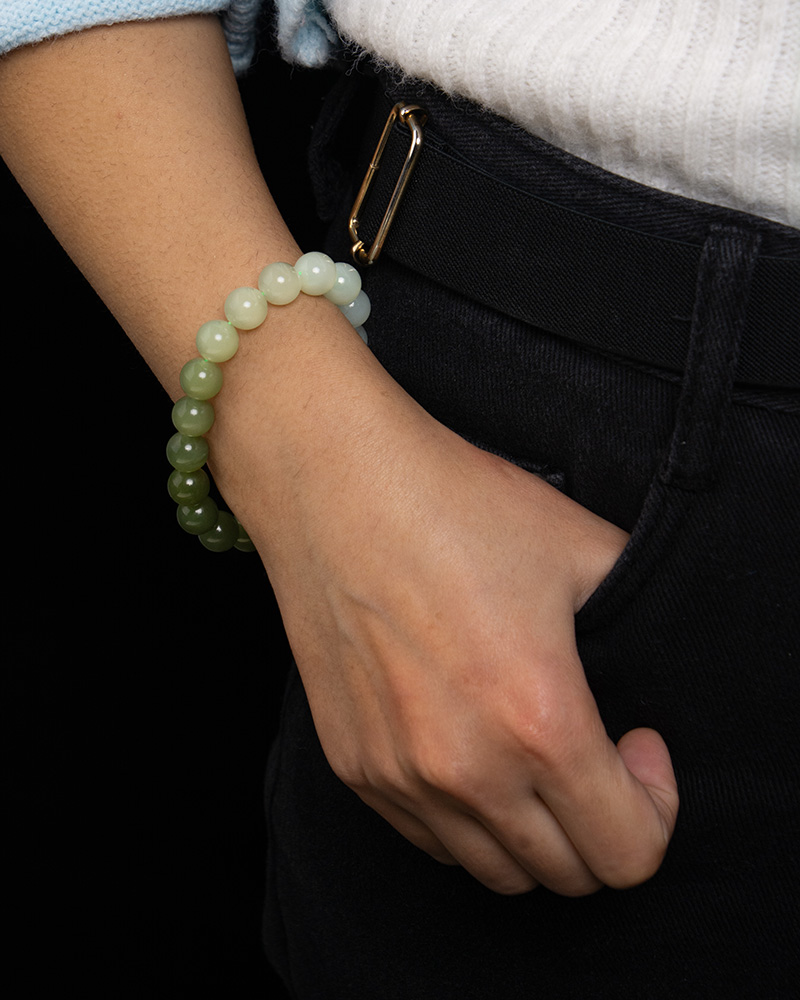 A person wears the Gradient Green Nephrite Jade Bracelet—celadon-to-olive green natural stone beads—resting their hand in a dark jeans pocket, with a white sweater at the wrist.
