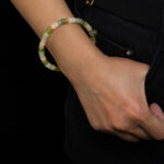 A person in black pants and a white shirt, right hand in pocket, showcases the Pastel Spectrum Nephrite Jade Bracelet – Heishi Rondelles with green, white, and beige beads on their wrist against a black background.