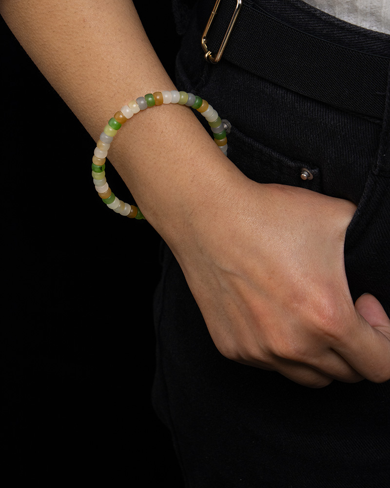LYU_4251 A person in black pants and a white shirt, right hand in pocket, showcases the Pastel Spectrum Nephrite Jade Bracelet – Heishi Rondelles with green, white, and beige beads on their wrist against a black background.