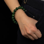 A person in dark pants and a white shirt wears the Deep Green Nephrite Jade Bracelet – 12mm Polished Round Beads on their wrist, with their hand resting casually at their hip against a black background.