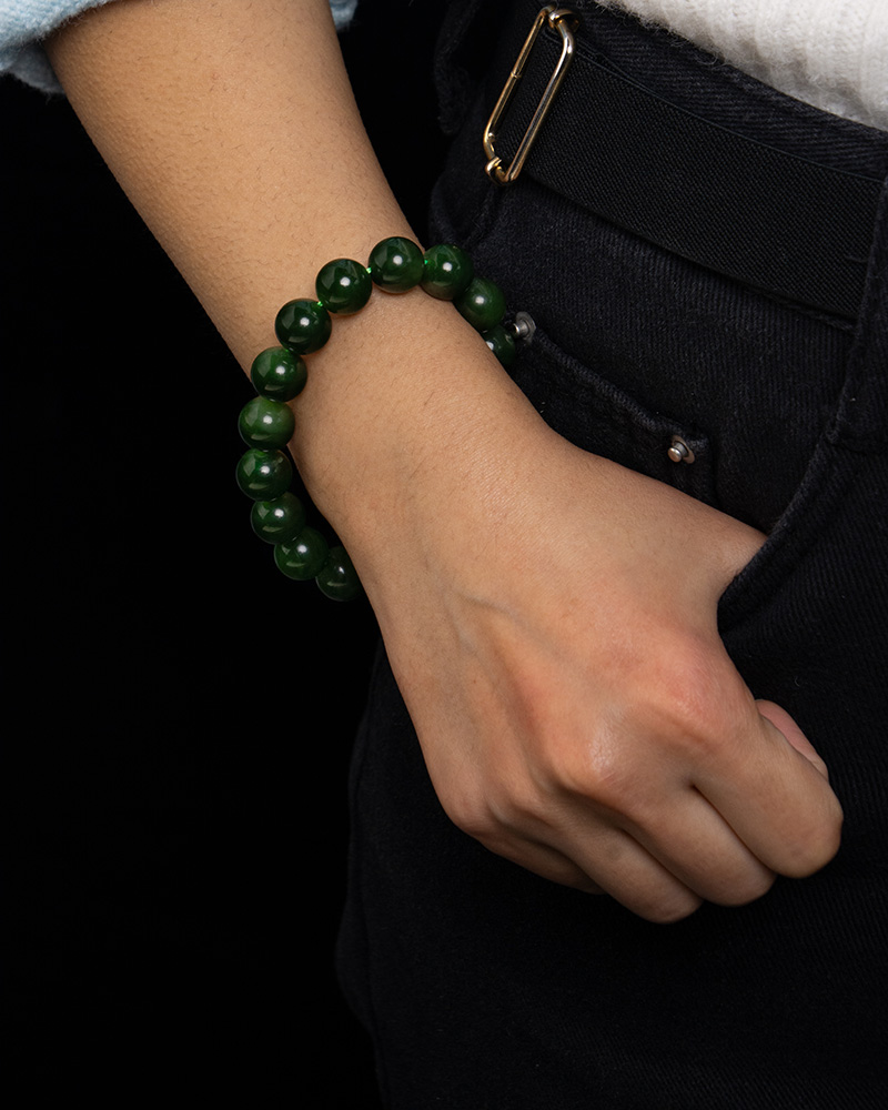 A person in dark pants and a white shirt wears the Deep Green Nephrite Jade Bracelet – 12mm Polished Round Beads on their wrist, with their hand resting casually at their hip against a black background.