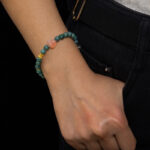 A person in black pants and a white top has their right hand in their pocket, showcasing the Green Jadeite Bracelet – round beads with a clear Buddha-head charm.