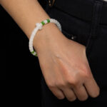 A person in dark pants and a white top rests their left hand near a pocket, showing off the Natural white Nephrite Jade Bracelet – Handmade Beads Stretch Bracelet with white and green beads and a cross-shaped accent.