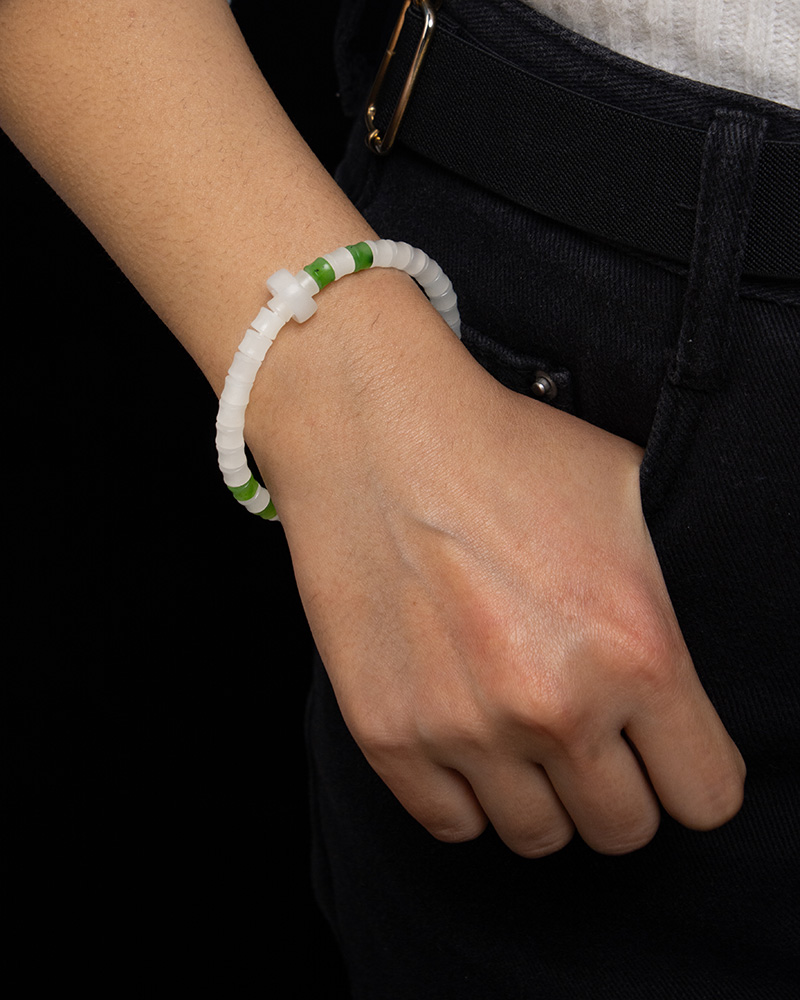 LYU_4258 A person in dark pants and a white top rests their left hand near a pocket, showing off the Natural white Nephrite Jade Bracelet – Handmade Beads Stretch Bracelet with white and green beads and a cross-shaped accent.
