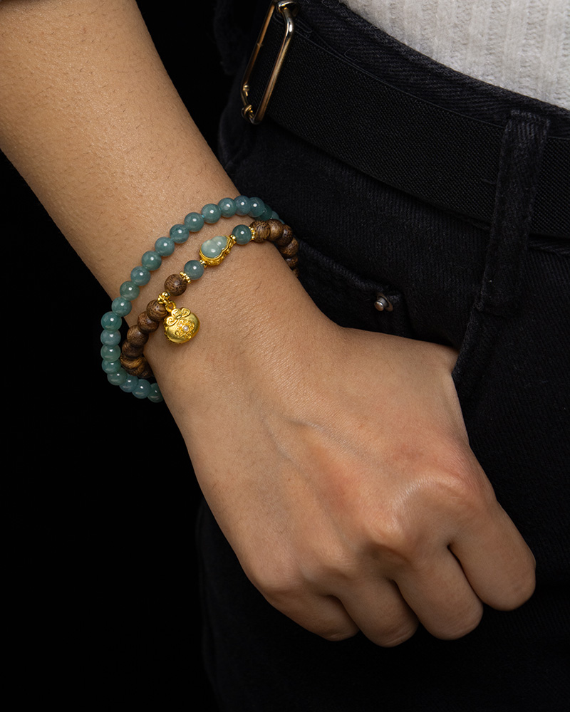 LYU_4260 A person in black pants and a white top wears the Sandalwood & Green Jadeite Double-Strand Bracelet with round beads and a pouch charm, shown on their wrist against a black background.