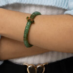 A person wearing the Nephrite Jade Bead Bracelet – Unique Ring with Natural Jade Skin, paired with a light blue sweater and black pants featuring a gold heart-shaped belt buckle, stands with arms crossed.