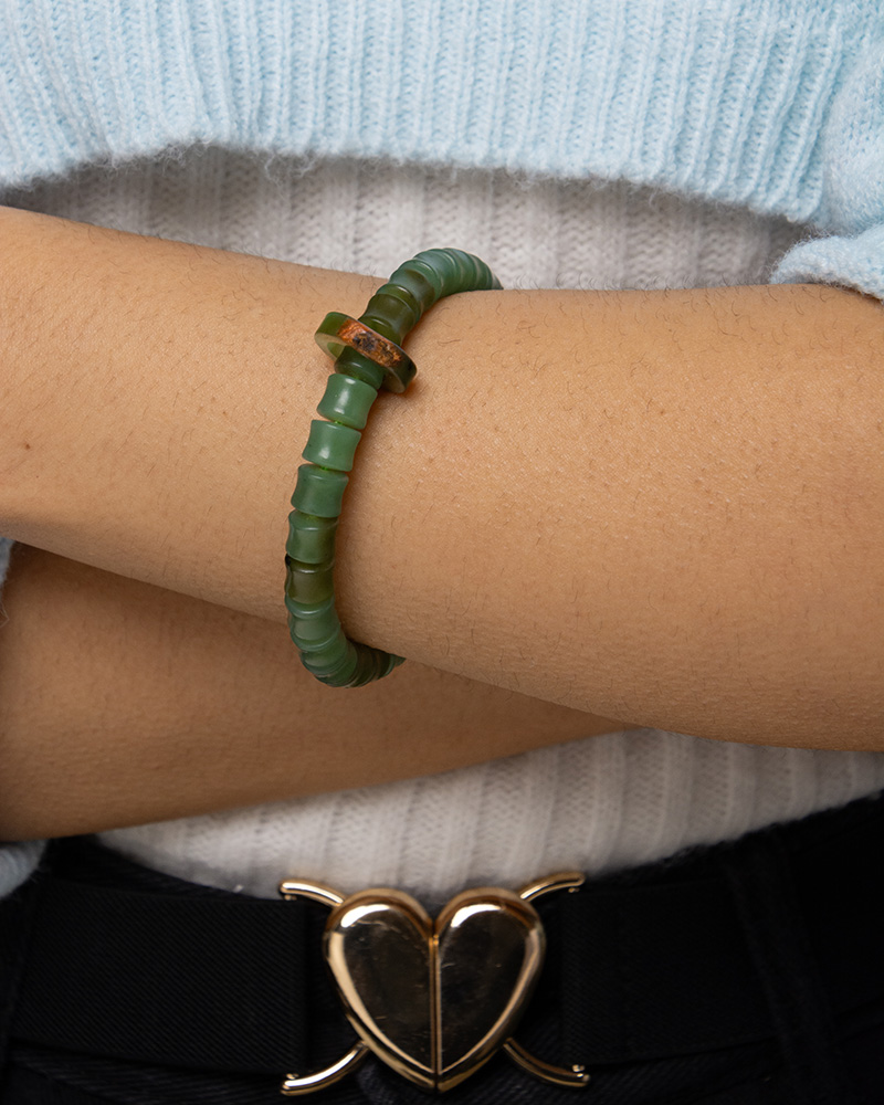 LYU_4287 A person wearing the Nephrite Jade Bead Bracelet – Unique Ring with Natural Jade Skin, paired with a light blue sweater and black pants featuring a gold heart-shaped belt buckle, stands with arms crossed.