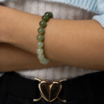 A person wearing a Gradient Green Nephrite Jade Bracelet with celadon-to-olive green natural stone beads and a light blue sweater crosses their arms. A gold heart-shaped belt buckle is visible above black pants.