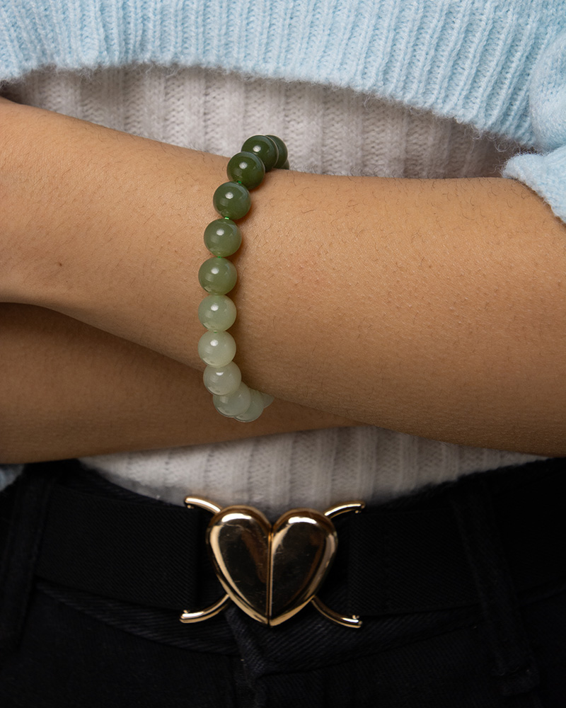 A person wearing a Gradient Green Nephrite Jade Bracelet with celadon-to-olive green natural stone beads and a light blue sweater crosses their arms. A gold heart-shaped belt buckle is visible above black pants.