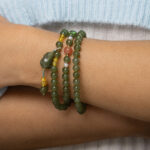A close-up of a wrist adorned with the Olive Green Nephrite Jade Wrap Bracelet featuring round beads, shown as multiple strands with yellow and reddish accents, set against a light blue and white fabric background.