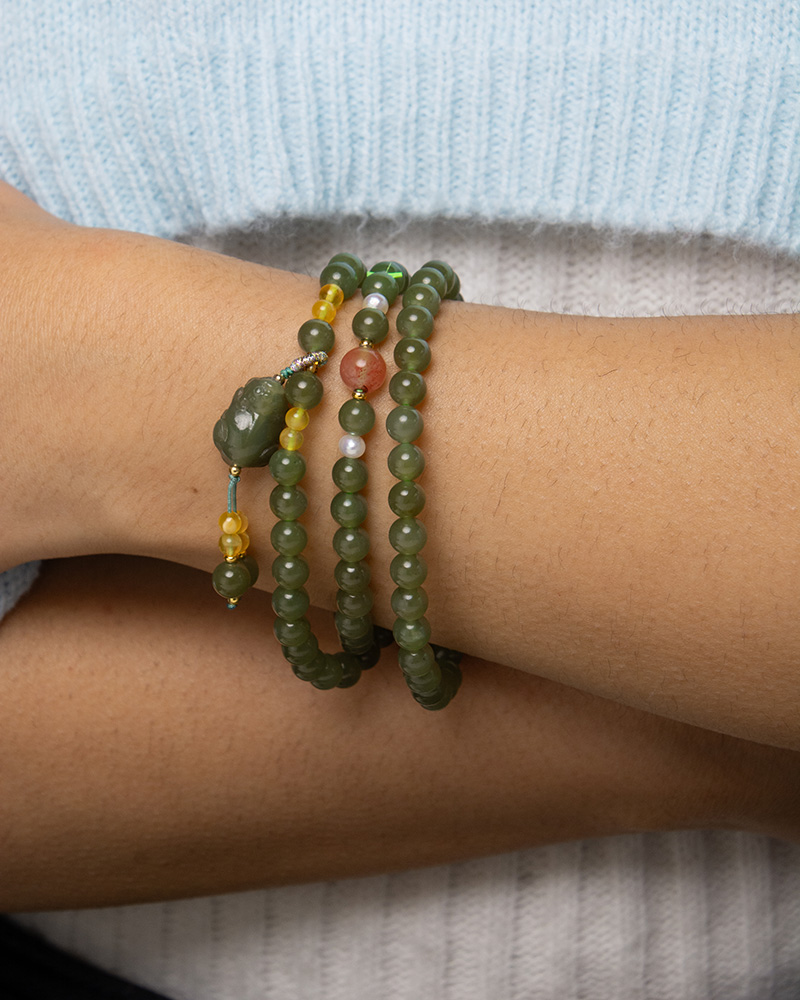 LYU_4291 A close-up of a wrist adorned with the Olive Green Nephrite Jade Wrap Bracelet featuring round beads, shown as multiple strands with yellow and reddish accents, set against a light blue and white fabric background.