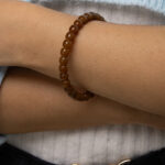 A person with arms crossed is wearing the Natural Hetian Jade Beaded Bracelet for meditation and energy. They are dressed in a white ribbed top, a light blue outer garment, and dark pants with a gold butterfly-shaped belt buckle.