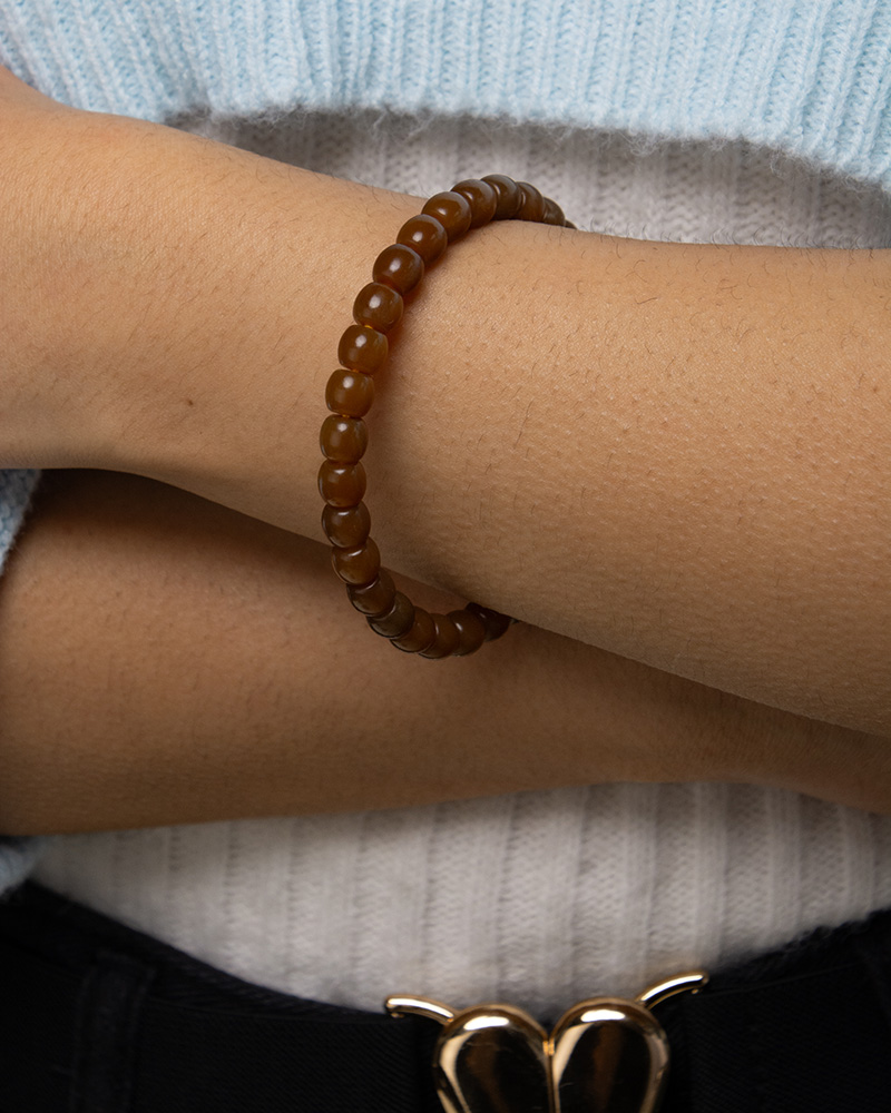 A person with arms crossed is wearing the Natural Hetian Jade Beaded Bracelet for meditation and energy. They are dressed in a white ribbed top, a light blue outer garment, and dark pants with a gold butterfly-shaped belt buckle.