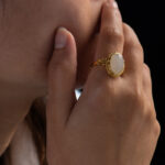 A person rests their hand on their chin, showcasing the Hetian White Nephrite Jade Ring—an oval cabochon set in a gold-tone filigree. The dark background highlights the ring’s elegant details and the wearer’s hand.