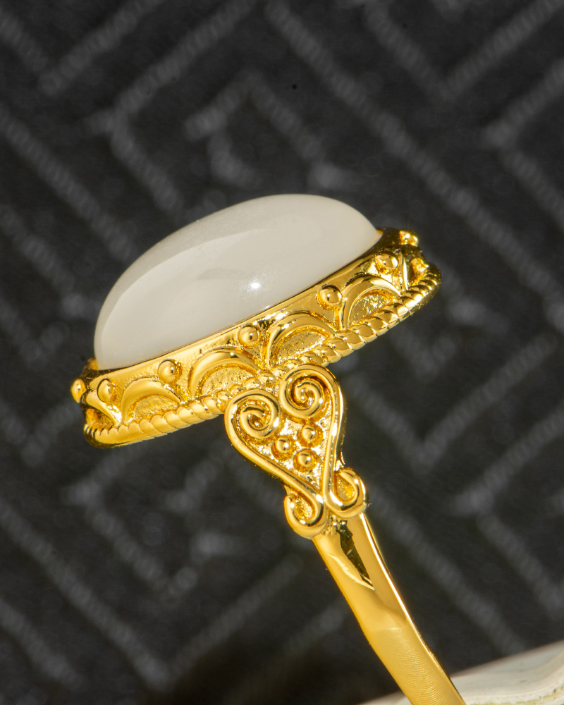 A close-up of the Hetian White Nephrite Jade Ring, showcasing an oval cabochon white jade set in an intricate gold-tone filigree setting against a dark, textured background.
