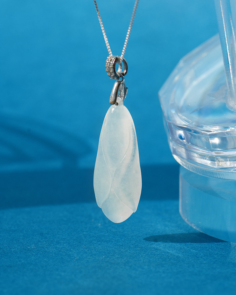 LYU_4493-1 A close-up of the Icy Green Jadeite Cicada Pendant with a hand-carved design and silver-tone bail, displayed on a silver chain against a blue background next to a clear, faceted object.