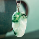 A close-up of the Jadeite Leaf Pendant, carved in green and white jadeite and suspended from a silver chain, with a blurred ceramic cup in the background.