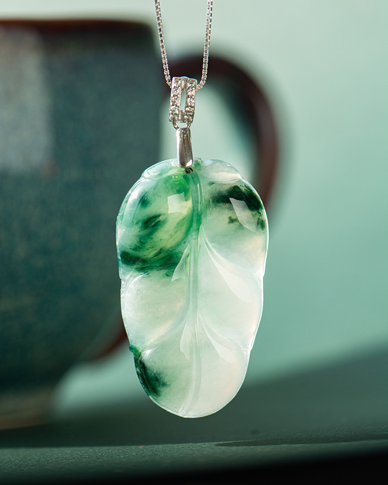 A close-up of the Jadeite Leaf Pendant, carved in green and white jadeite and suspended from a silver chain, with a blurred ceramic cup in the background.
