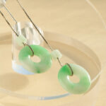 Light Green Jadeite Donut Drop Earrings feature circular jadeite pendants with central holes on silver hooks, elegantly displayed on a clear round stand against a softly lit beige background.