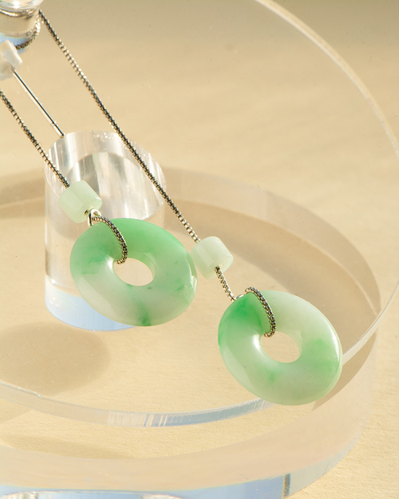 Light Green Jadeite Donut Drop Earrings feature circular jadeite pendants with central holes on silver hooks, elegantly displayed on a clear round stand against a softly lit beige background.