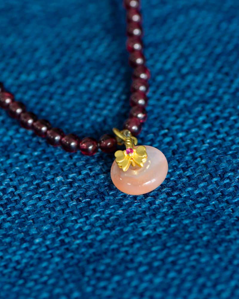 A close-up of the Wine-Red Garnet Wrap Bracelet, featuring round dark red beads and a persimmon charm pendant, displayed on textured blue fabric.