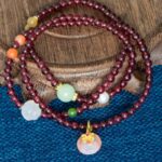 The Wine-Red Garnet Wrap Bracelet, featuring round beads and a persimmon charm, is coiled on a wooden surface above blue textured fabric.