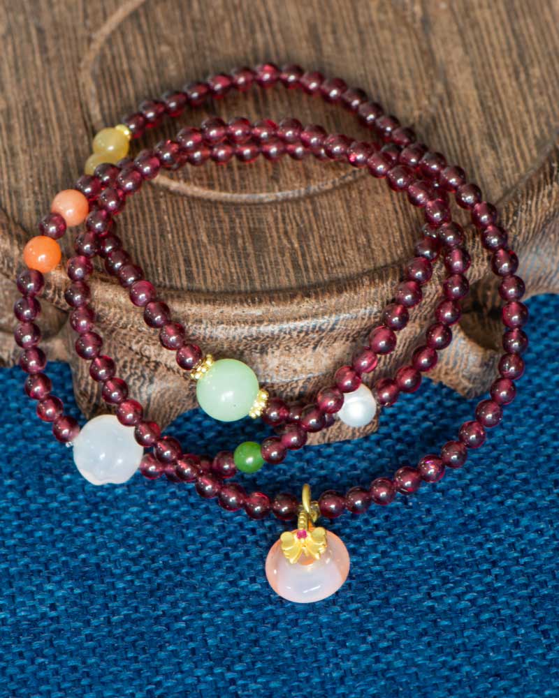 The Wine-Red Garnet Wrap Bracelet, featuring round beads and a persimmon charm, is coiled on a wooden surface above blue textured fabric.
