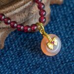 The Wine-Red Garnet Wrap Bracelet features round wine-red beads and a persimmon-shaped charm, showcased on textured blue fabric beside a wooden surface.