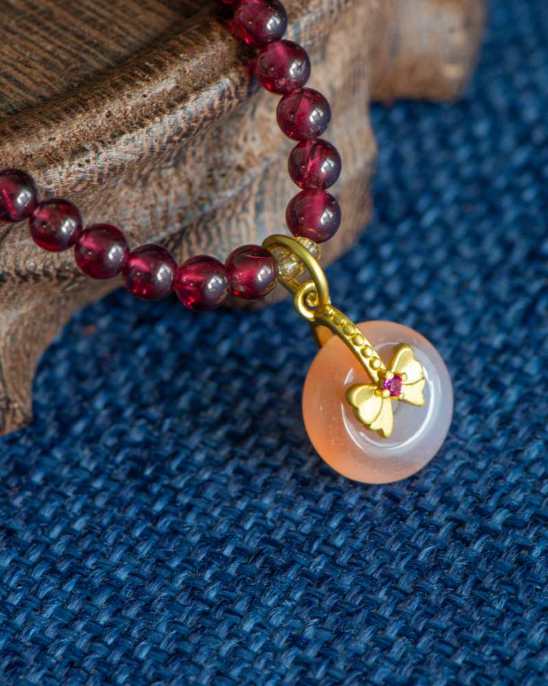 The Wine-Red Garnet Wrap Bracelet features round wine-red beads and a persimmon-shaped charm, showcased on textured blue fabric beside a wooden surface.