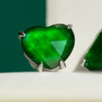 Close-up of a Vivid Green Jadeite heart-cut stud earring with custom faceting, set in 925 sterling silver; blurred green and white background.