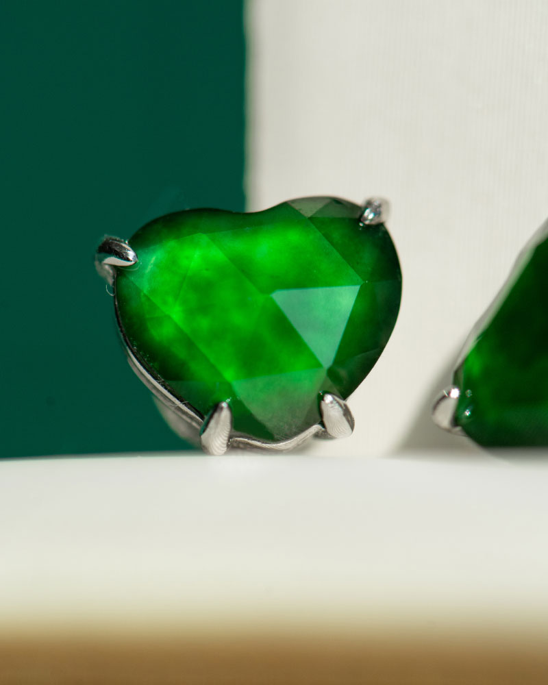 Close-up of a Vivid Green Jadeite heart-cut stud earring with custom faceting, set in 925 sterling silver; blurred green and white background.