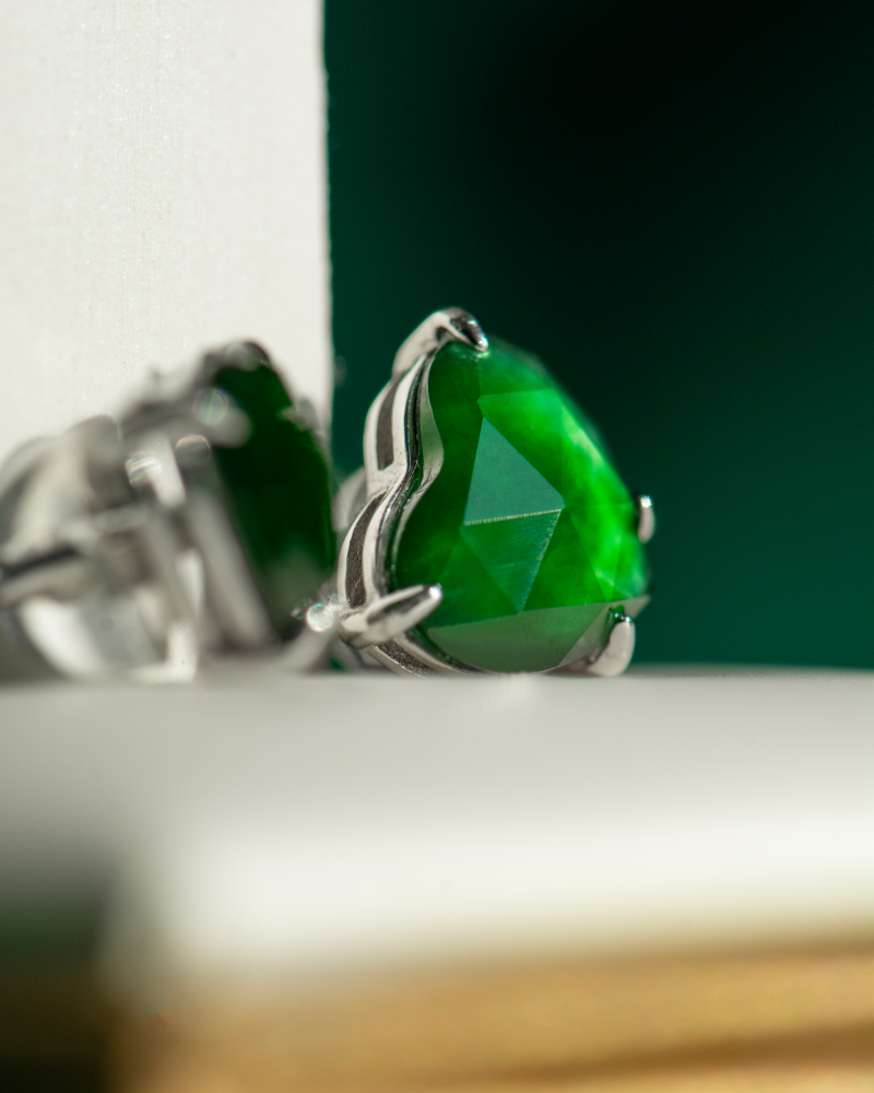 Close-up of Vivid Green Jadeite Stud Earrings, heart-cut with custom faceting, set in 925 sterling silver, shown against a blurred white and green background.