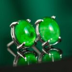 A pair of Vivid Green Jadeite Stud Earrings with round cabochon gemstones set in 925 sterling silver, displayed on a green background with visible reflections.